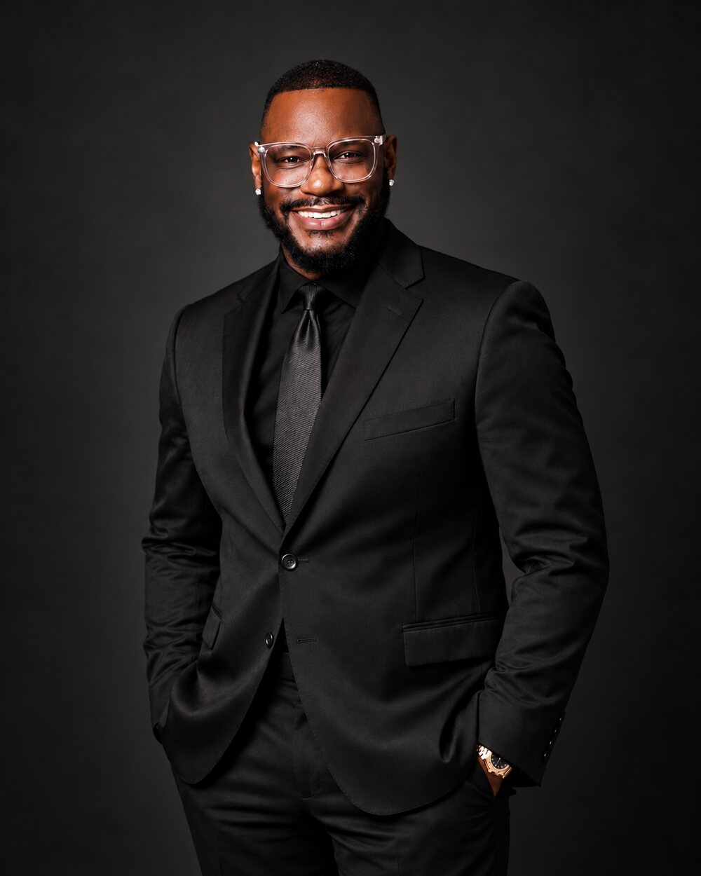 Ortavia Simon standing in a black suit and tie against a dark studio backdrop, hands in pockets